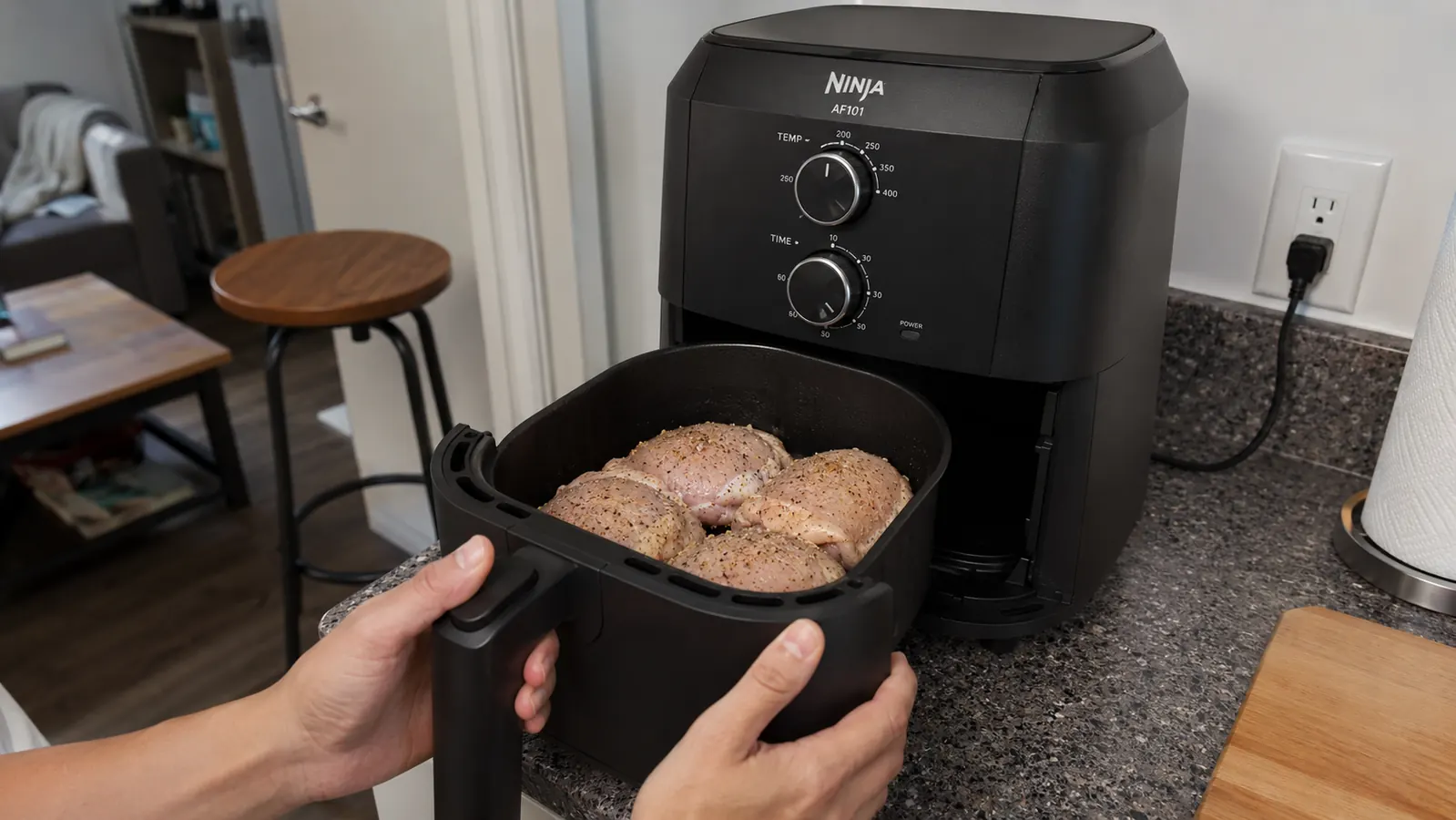 Hand placing a basket of seasoned chicken thighs into the Ninja AF101 air fryer on a small counter